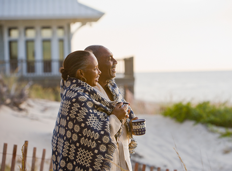 An older couple wrapped up in a blanket smile, looking out over the ocean.